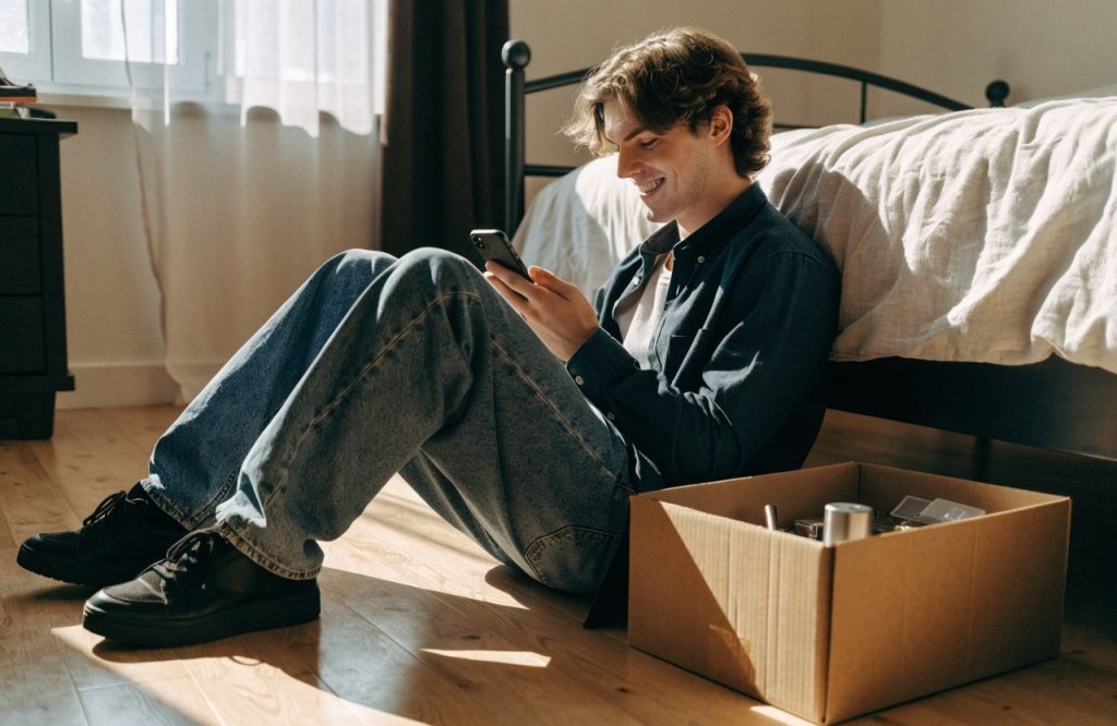 Rapaz sentado no chão do quarto sorrindo para o celular, com uma caixa aberta ao lado